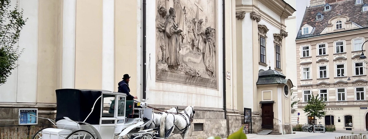The image shows a traditional horse-drawn carriage (fiaker) passing by the exterior wall and large religious relief mural of St. Peter's Church (Peterskirche) in Vienna, Austria